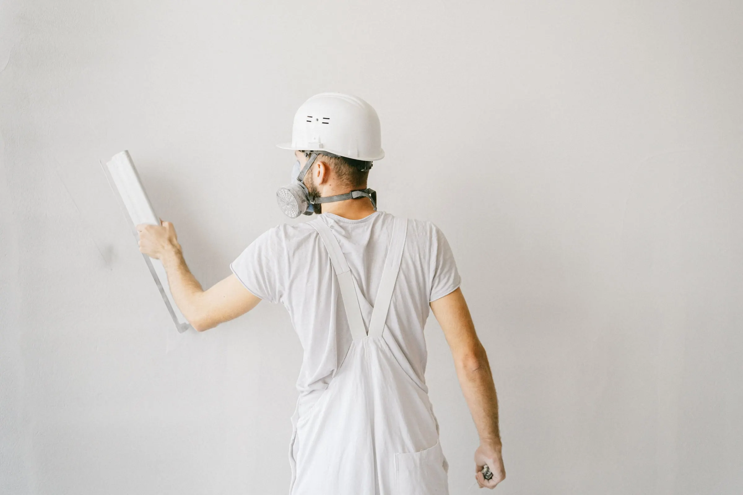 Tradesman in a hard hat and respirator smoothing a freshly plastered white wall with a straightedge