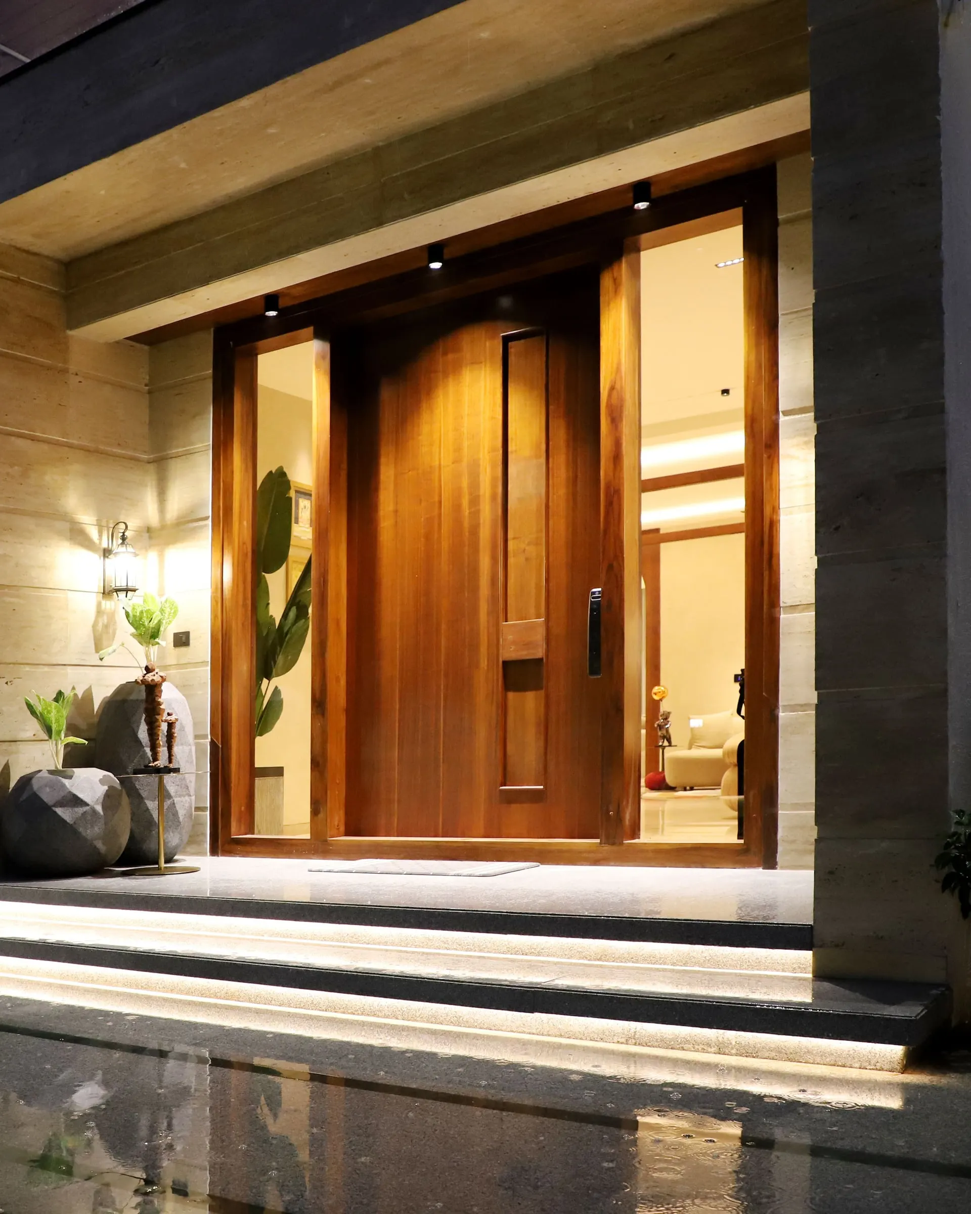 Warmly lit entrance of a modern home at night, with a tall timber front door, stone-clad walls and lit steps